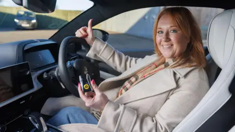 Omaze Claire is wear sat in the driver's seat of the car holding up a set of car keys with one hand and thumbs up in the other and is smiling at the camera.