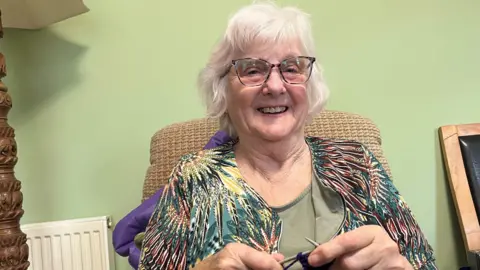 John Devine/BBC Adoree Webb has grey permed hair and tortoiseshell framed glasses. She has a colourful cardigan on over a light green top, behind her is a light green wall.