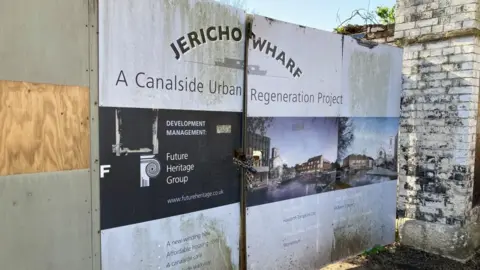 A gate is boarded up with a sign saying Jericho Wharf, a canalside urban regeneration project. It looks old and run down.