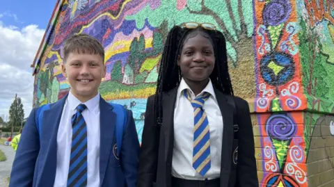 A young boy in a white shirt, blue and black tie and blue blazer and a girl with a black blazer, white shirt, blue and yellow tie and black hair both smiling at the camera. You can see a colourful mural on the side of a wall behind them depicting a scene in nature.