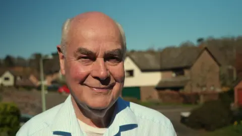 Shaun Whitmore/BBC A head shot of Mark Packard. He is smiling and wearing a striped light blue short over a white T-shirt. He is looking into the camera. 