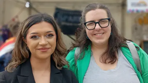 Two women are standing together and smiling at the camera. On the left is a young woman with tan skin and brown hair with blond highlights. On the right is a woman with dark hair and glasses.