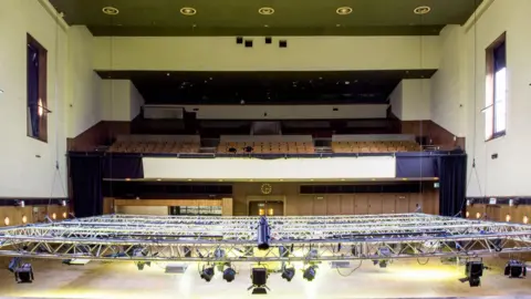 Ian Grundy The interior of Borough Hall showing an empty auditorium viewed from the stage, with lighting rigging suspended above the floor, rows of seating on two levels and a large balcony at the rear.