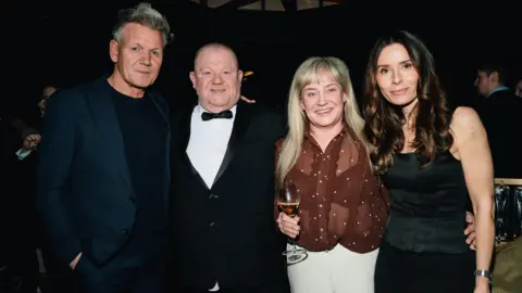 Gordon Ramsay on the left stood next to a man wearing a suit and a black bow tie, next to a woman wearing a burgundy blouse and holding a wine glass who is standing next to a woman with brown hair.