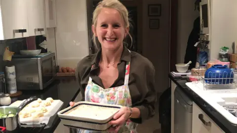 Abinger Cookery School Lizzie Marsh standing in her kitchen wearing a brown shirt and apron, holding a ceramic dish and smiling at the camera