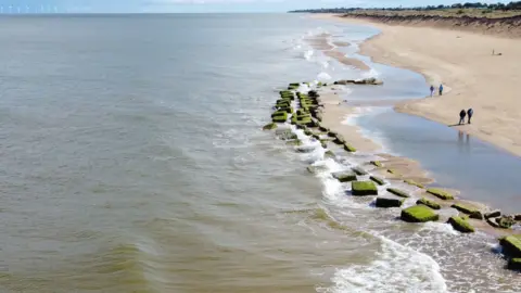 John Fairhall/BBC An aerial shot of a row of seaweed covered concrete anti-tank blocks emerging out of the sea at Winterton. In front to the left is the grey sea, with blue sky above and in the far distance wind turbines. To the right there is a sandy beach, with a handful of people walking along it, and sandy dunes rising to a low cliff. 