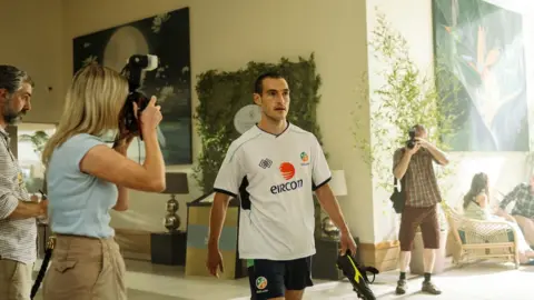Aidan Monaghan Éanna Hardwicke playing Roy Keane, who is walking through a hotel lobby in a white away Republic of Ireland top, having his photo taken from either side by a man and a woman. 