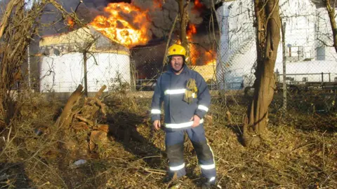 David Humphreys A firefighter stands in front of a large industrial fire. Behind the firefighter, two white storage tanks are partially engulfed in flames.