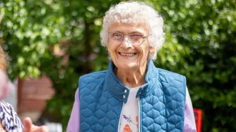 Pam Haylip smiles at the camera as she stands in a garden. She is wearing a blue gilet with a purple cardigan, and white T-shirt with a flower pattern on the front. She has short grey hair and is wearing glasses. 