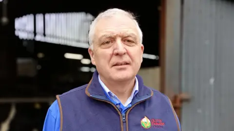 William looking at the camera. He is wearing a blue long sleeve top with a navy fleece body warmer over it.