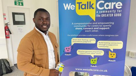 A man stands beside a pop up banner displaying information about ‘We Talk Care’