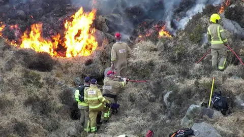 The picture shows five firefighters in tan coloured uniforms tackling a fire on the gorse covered slope of a mountain. Flames, smoke and blackened vegetation are visible above them.   