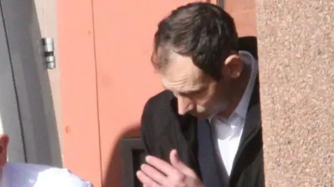 Adam McManus being led away from court, he is wearing a dark jacket and a white shirt, and has his head lowered