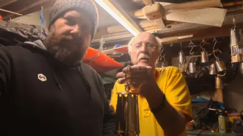 Supplied A man with short, white hair, a white moustache, glasses, a yellow polo shirt and dark trousers is holding a miners lamp. He is standing in a shed filled with other mining lamps and tools. He is passing the lamp to another man with a thick dark beard, black wooly hat and black hoodie. Strip lighting illuminates the shed.
