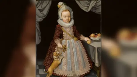 A portrait of a child dressed in a long dress-like gown with a lace neck ruff, hat and lace apron. The child has a gold chain over his shoulder and is holding a rattle with one hand and an apple with the other. There is a small dog jumping up at his side.