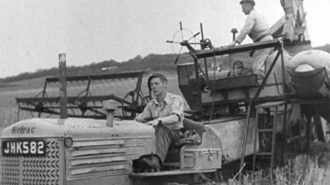 Black and white picture.  Combine harvester in a field, with man with cap driving and another standing on the back.  A small boy also sits behind.