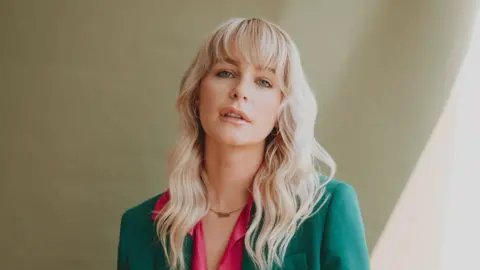 Rhiannon Holland Jess Davies, wearing a green suit jacket and pink shirt, looks directly at the camera in a headshot.