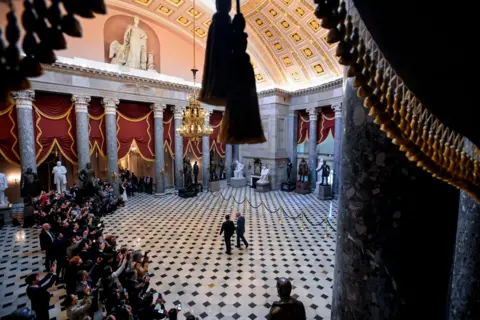AP A wide view shows the King being led through the hall of the Capitol with reporters to the side.