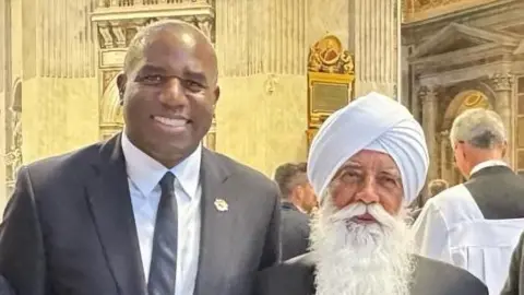 Bhai Sahib A man with close-shaved grey hair, wearing a dark suit with a white shirt and a dark tie. Next to him is the bearded man from the above pictures.