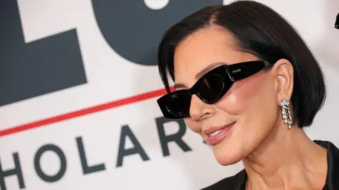 Kris Jenner, wearing a pair of thick black sunglasses and dangling earrings, smiles for photographers at a red carpet event.