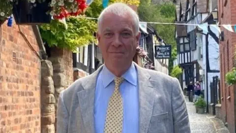 Ewen Sinclair is standing in a street in Ledbury, wearing a suit and tie - behind him are several black and white buildings.
