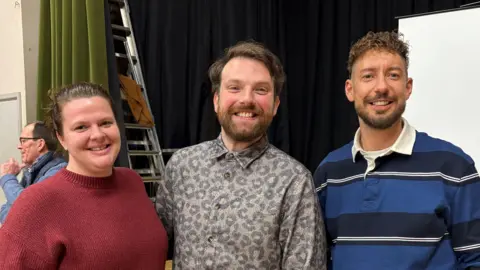 Louis Reynolds/BBC Image showing Chloe Peacock, Steven Cameron and Charlie Aldridge stood in front of the stage at Great Ouseburn Village Hall