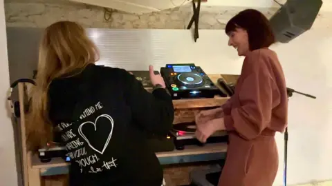Milly with a course participant laughing and smiling. They are standing in front of a set of DJ decks, facing away from the camera. The participant, who has long blonde hair and a dark hoodie on, is doing a thumbs up sign to Milly who is on the right.