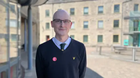 A bald man in glasses, wearing a V-neck jumper, a shirt and tie. He is standing outside a large brick building which looks like a former mill.