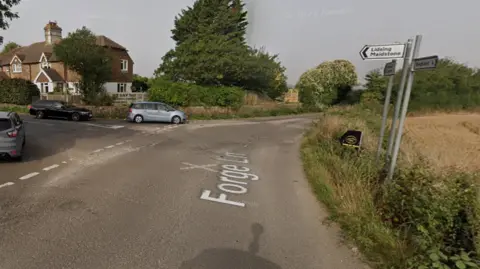 A Google Map image of the road into Lidsing, on the outskirts of Maidstone, Kent. There are three cars to the left and a field to the right. 