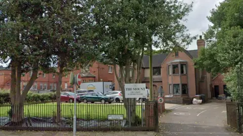 Several red coloured brick buildings which include Victorian and more modern buildings. The buildings have long windows and one chimney is in shot and there are parked cars and a wide driveway. There is also a green lawn and a white sign which says 'The Mount' at the front of the picture. 