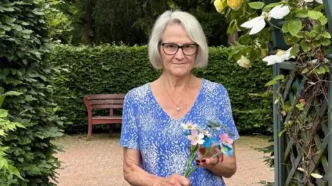St Richard's Hospice A woman with a grey bob and black glasses wears a blue top with white pattern and v-neck. She holds a bunch of metal flowers that are pink, white and blue, and stands in a paved garden with a hedge and bench behind.