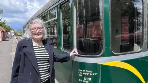A woman stands on a railway platform with one hand resting on a green vintage rail carriage, with station buildings and tracks visible behind them under a cloudy sky.