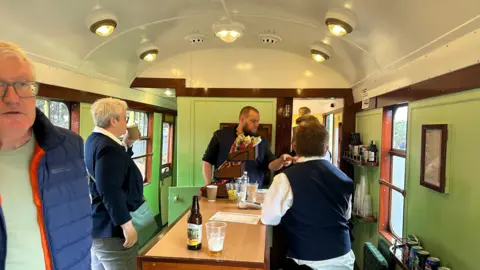 A bar area is painted in green and there is a framed sign on the wall. A woman is serving drinks. People are standing around and talking. 