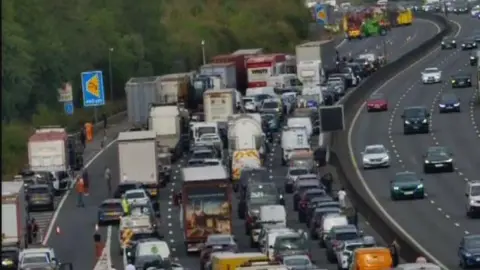 National Highways Long queues of motorway traffic, with people getting out of their stationary vehicles to stand on the carriageway. 