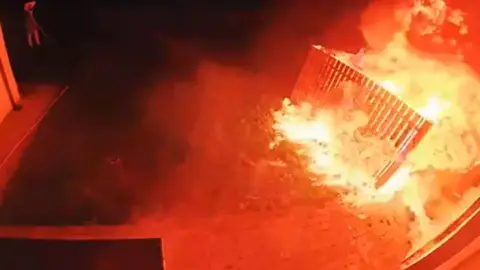 Victoria Nichols Doorbell footage showing the fire - the blaze is to the right of the image and is engulfing a wooden fence. To the top left a person can be seen with their arms on their head. The image is mostly red and orange due to the light from the fire, with some black to the left.