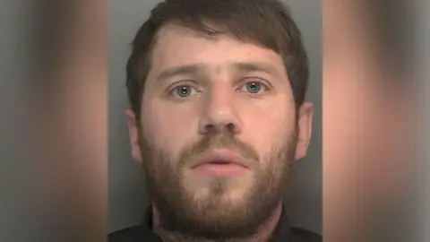Merseyside Police A mugshot of a man with short dark hair and blue eyes, wearing dark clothing on a grey background