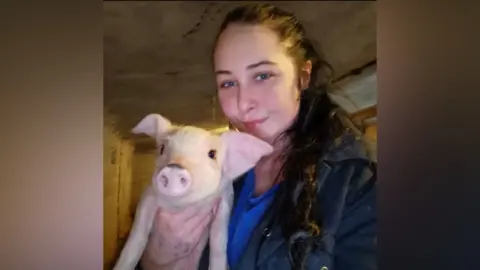 Contributed A woman with dark hair which has been tied back and the ponytail rests over her shoulder takes a selfie as she holds a small piglet. She is wearing a navy coat with a lighter blue top underneath. 