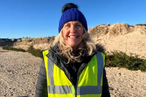 Carolle Ralph, woman with blonde hair smiling at camera, wearing hi-vis yellow jacket and warm blue hat, on a sandy beach with Christmas trees lying on the ground in front of dunes.