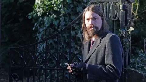 Benjamin Clarke outside Durham Crown Court. He has long dark hair and a beard, and is wearing a dark coat, black gloves and a dark red tie. He is looking into the camera and smoking a cigarette while holding a phone.