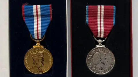 John Huggins A gold medal featuring Queen Elizabeth's head and wording with blue, white and burgundy striped ribbon. Next to it is a matching medal in silver with a different striped ribbon but in the same colours. They are both on a black background.