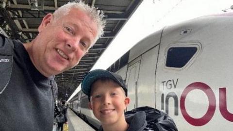 Dave Chalk and his son Viktor take a selfie in front of a train as they smile at the camera