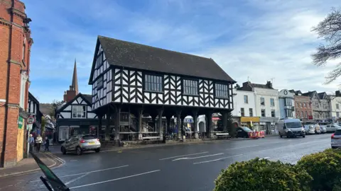 A wide street scene in a historic town, centred on a large black-and-white timber-framed market hall raised on wooden pillars.