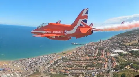 A small red jet with Union Flag livery on its tail flies over a coastal town, with the sea seen to the left of the image. The picture is taken from the cockpit of another plane flying alongside it.