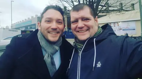 Martin Hywood (L-R) Jon Richardson and Martin Hywood. 
Jon is wearing a black coat and a grey scarf and smiling at the camera.
Martin is waring a navy jacket and a scarf while holding the camera in order to take the selfie.
They are outside and the sky is looking cloudy and the trees are bare of any leaves