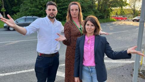 Three councillors raising their hands in disapproval as they stand by a partially closed roundabout on a summer's evening