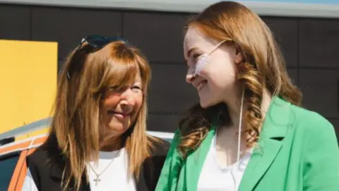 Debbie Roscoe Debbie has long brown hair and a white T'shirt on with a black vest over the top. Her daughter also has long brown hair and is wearing a green jacket over a white top. She has a tube coming out of her nose that is taped to her face and runs down the side of her neck. The pair are looking at one another and smiling.