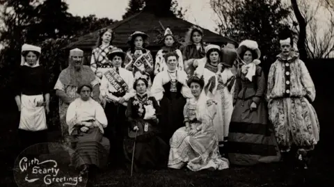 Paul Andersen A black-and-white photo, possibly from the early 20th Century, of a group of 14 men and women wearing various fancy dress costumes, including a clown suit, knight's outfit and elaborate dresses.