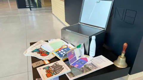 The picture shows the capsule open and items around it which will be put inside to be buried. They include a copy of the local paper, a water bottle, pictures drawn by children, a book on art, a train ticket, and a bag.