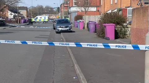 Nikki Robertson/BBC Police tape can be seen in the foreground, tied across a residential street. Pink and purple bins line the side of the pavement. In the background there is a police van and a group of officers are standing in front of it. 