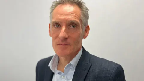 A white man called Nick Hynes. He has short grey hair which looks slightly ruffled. He is wearing a grey suit jacket and a grey shirt.